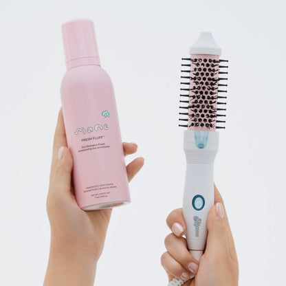 Hand holding a pink bottle labeled 'Fresh Fluff' and a white hairbrush with black bristle head on a light gray background.