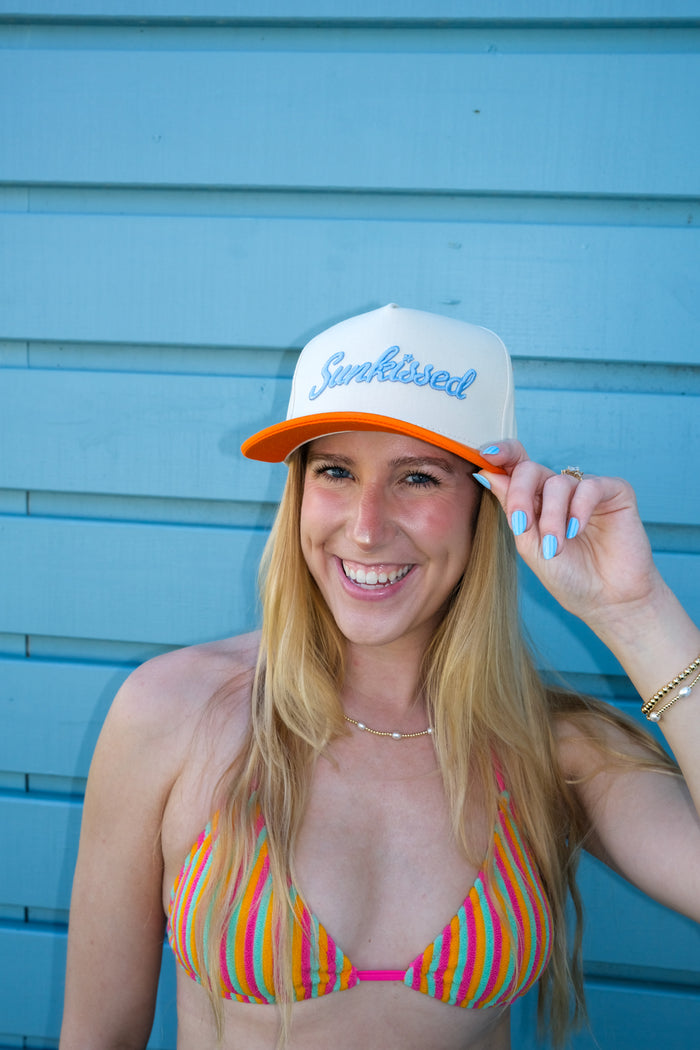 Woman wearing a colorful striped bikini and 'Sunkissed' cap against a blue wall